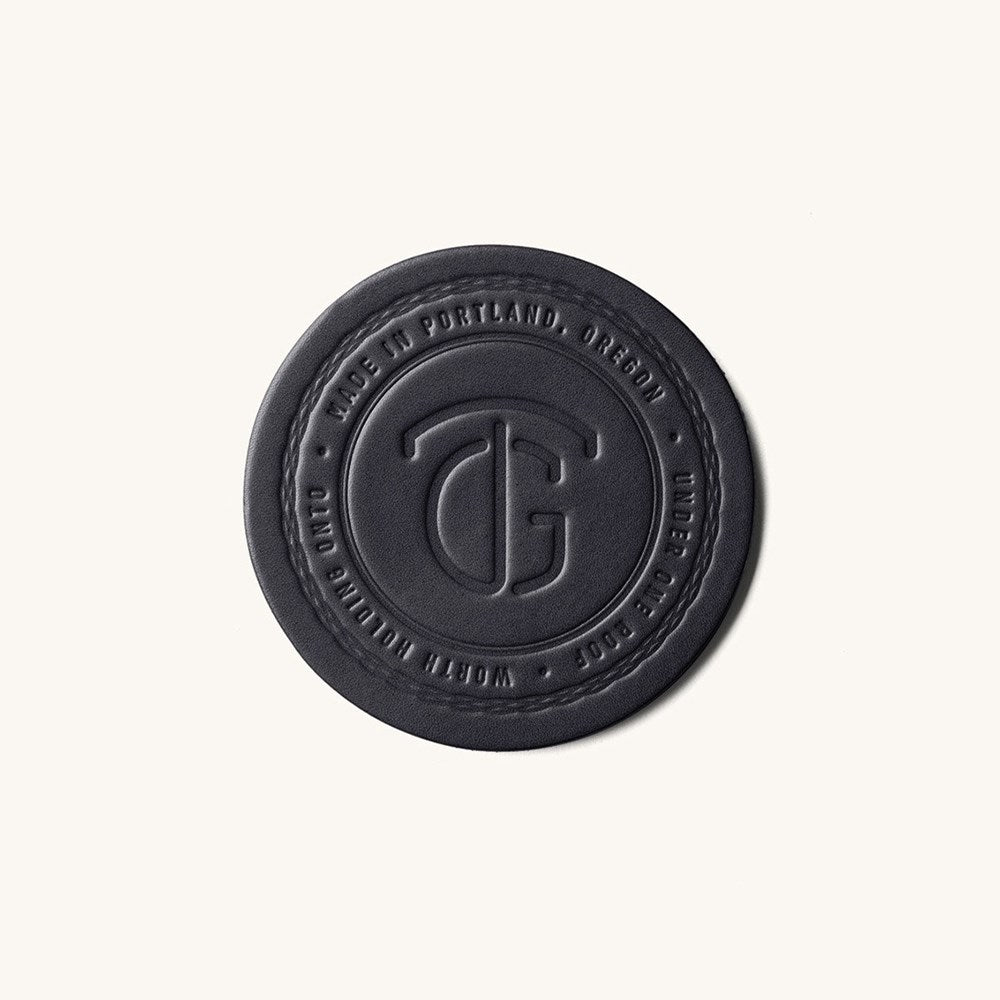 Black round coaster by TG, embossed with a stylized logo and "MADE IN PORTLAND, OREGON" text, with a stitched edge.