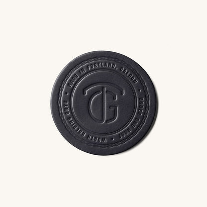 Black round coaster by TG, embossed with a stylized logo and "MADE IN PORTLAND, OREGON" text, with a stitched edge.