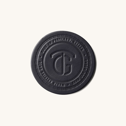 Black round coaster by TG, embossed with a stylized logo and "MADE IN PORTLAND, OREGON" text, with a stitched edge.