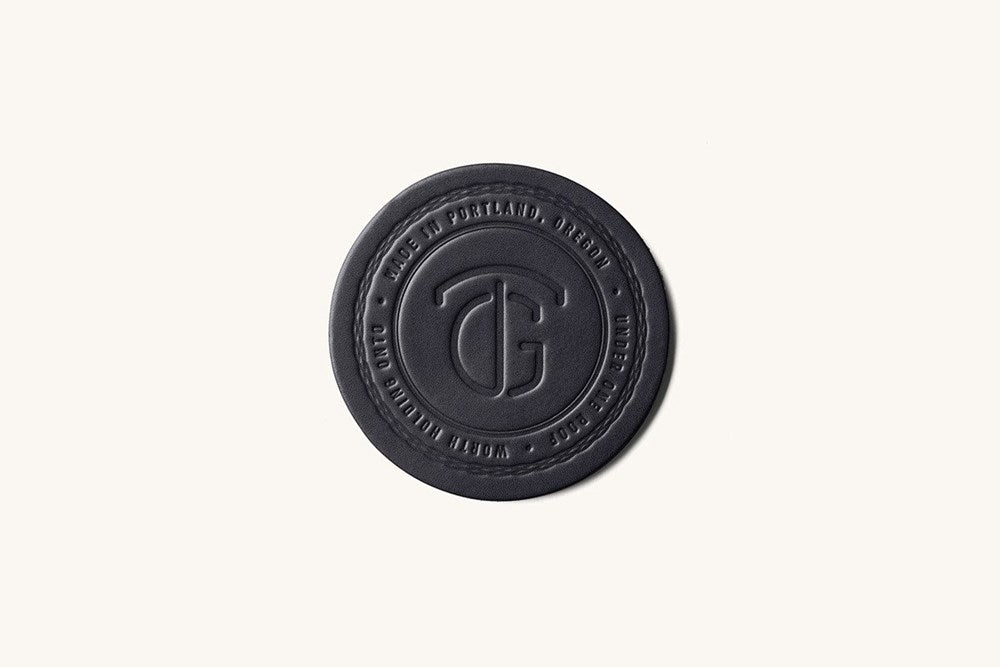 Black leather-like round coaster by TG, featuring a debossed logo and circular text details.