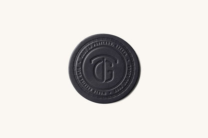 Black leather-like round coaster by TG, featuring a debossed logo and circular text details.