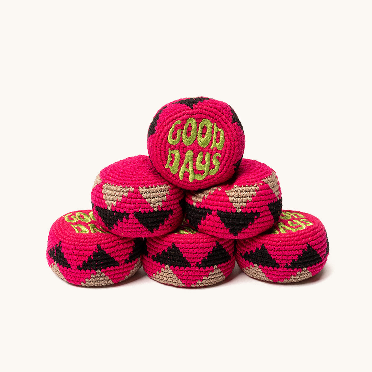 Six fuchsia knitted footbags with black and beige geometric patterns, stacked in a pyramid, one embroidered with 'GOOD DAYS'.