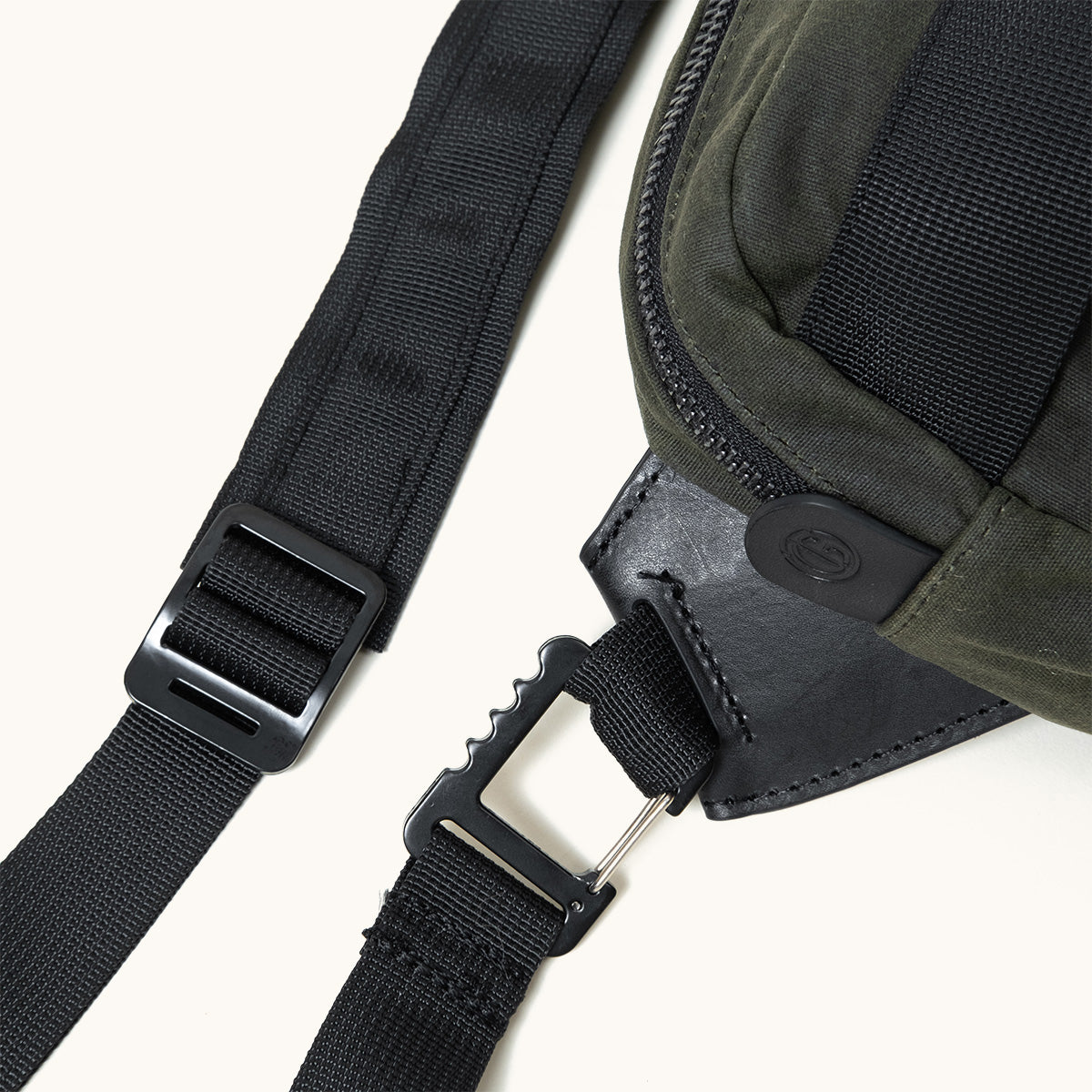 Cropped view of a juniper green waxed canvas crossbody pack with black zipper, leather trim, and nylon straps with black plastic hardware.