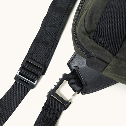 Cropped view of a juniper green waxed canvas crossbody pack with black zipper, leather trim, and nylon straps with black plastic hardware.