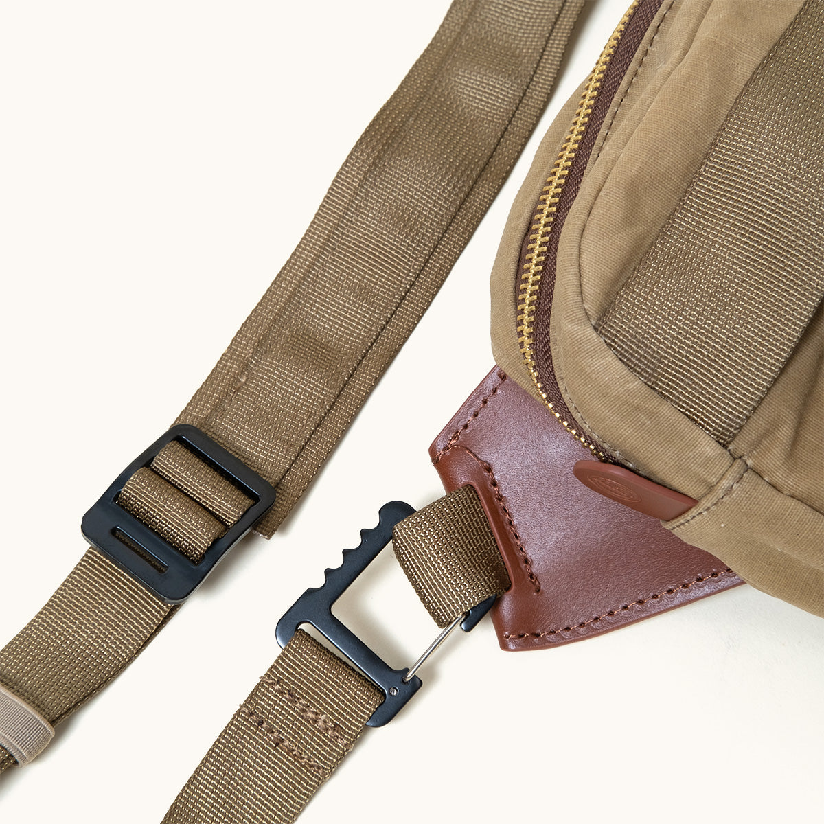 Utility tan waxed canvas crossbody pack with a brown leather accent, brass zipper, and black buckles on olive green webbing straps.