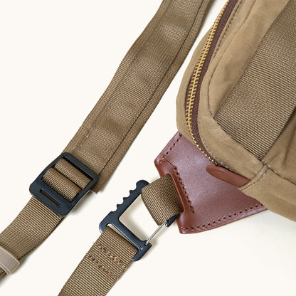 Utility tan waxed canvas crossbody pack with a brown leather accent, brass zipper, and black buckles on olive green webbing straps.