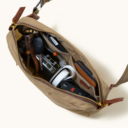 Utility Tan waxed canvas crossbody pack, open from overhead, revealing an organized interior with keys, a wallet, and a water bottle.