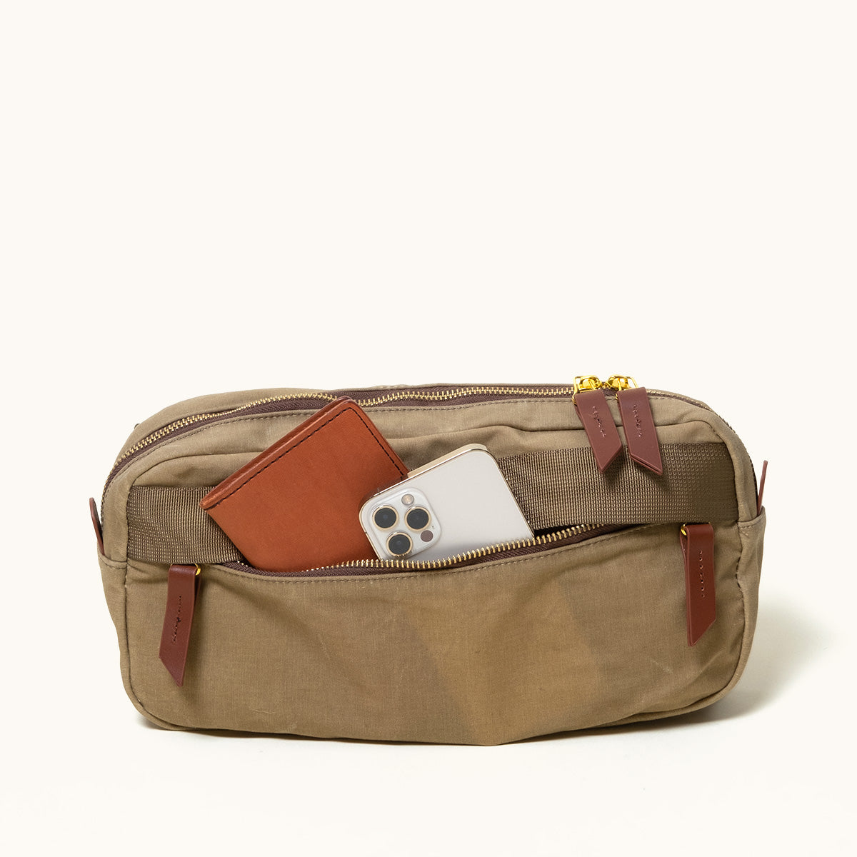 Utility Tan waxed canvas crossbody pack with an open front pocket showing a brown wallet and a silver smartphone, featuring brass zippers and leather pulls.