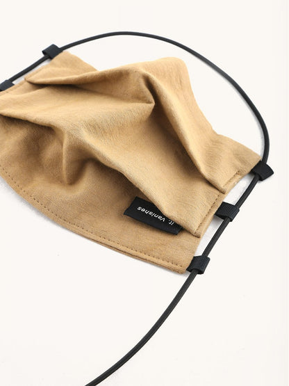 Tan fabric face covering with a small black "It. Vanishes" label and black elastic straps with adjusters.