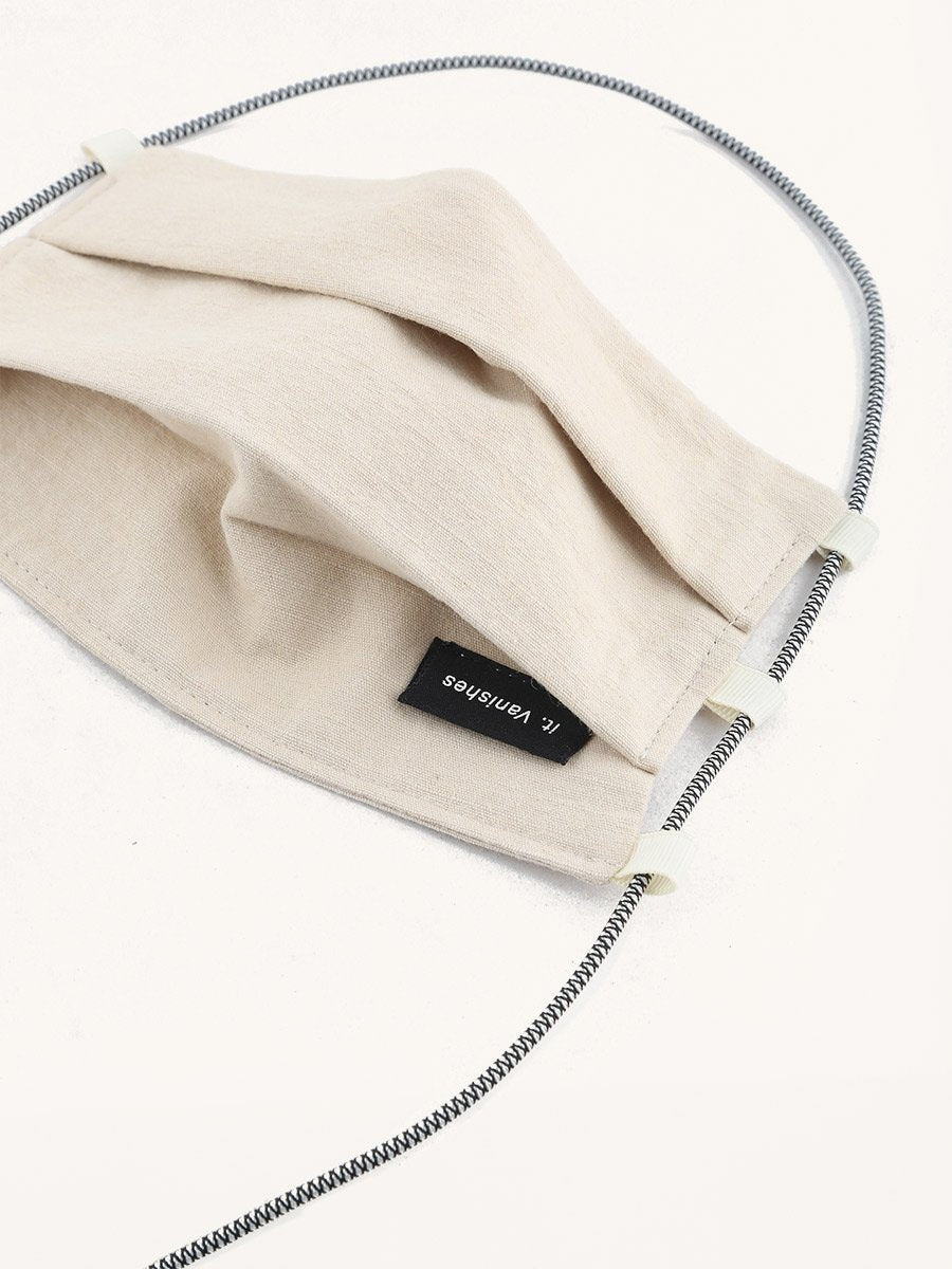 Beige pleated fabric face covering with a small black label featuring 'it. Vanishes' text, white strap adjusters, and a striped black and white elastic strap.