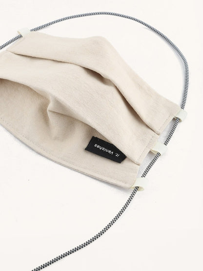 Beige pleated fabric face covering with a small black label featuring 'it. Vanishes' text, white strap adjusters, and a striped black and white elastic strap.
