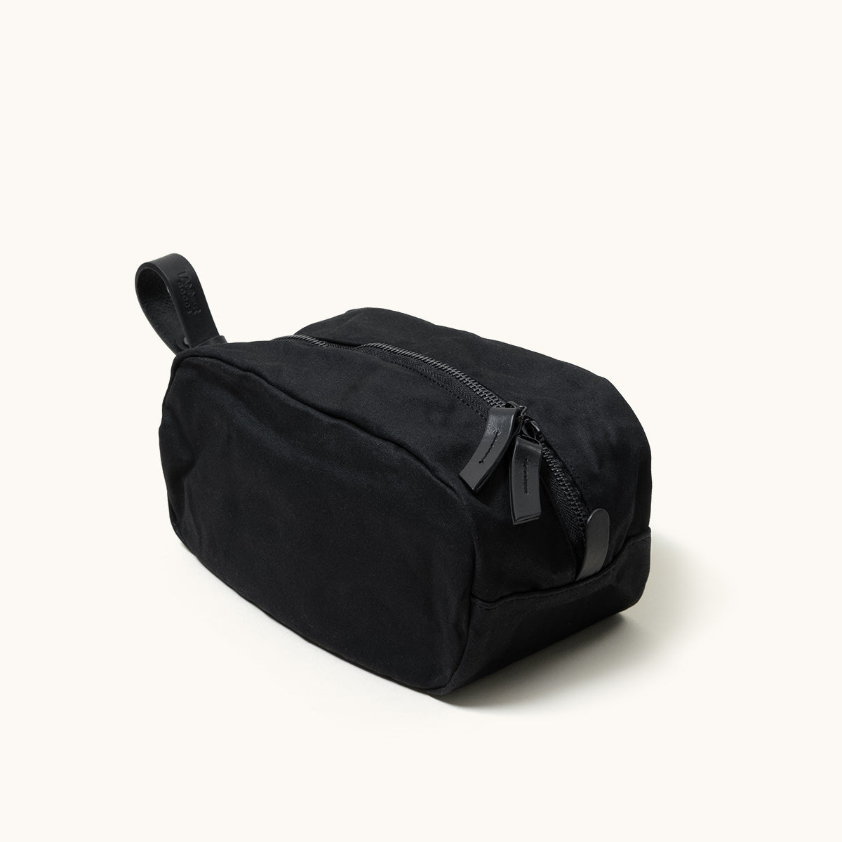 Black canvas Dopp Kit by Drifter with a top zipper, two leather pulls, and a leather carry handle, shown at an angle.