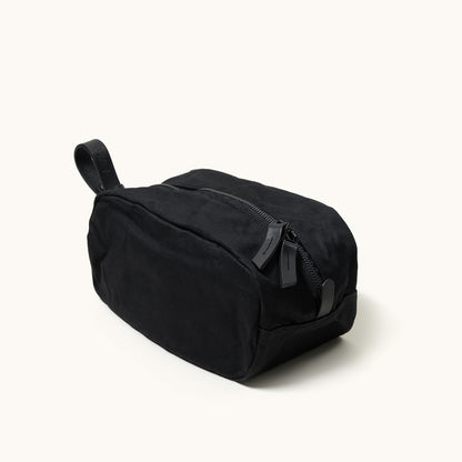 Black canvas Dopp Kit by Drifter with a top zipper, two leather pulls, and a leather carry handle, shown at an angle.