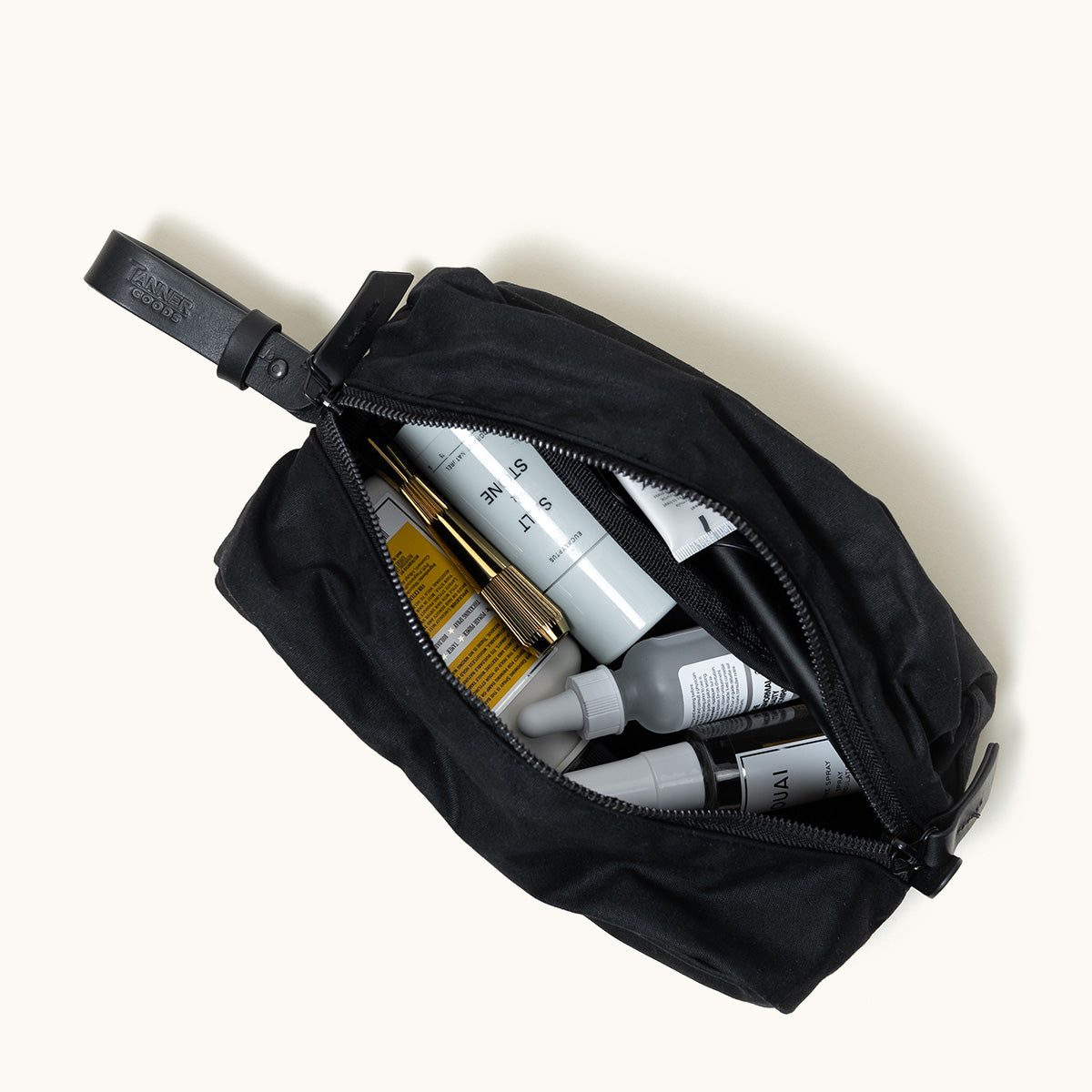 Black waxed canvas Dopp Kit by Tanner Goods, open with toiletries inside, angled overhead on a white background.
