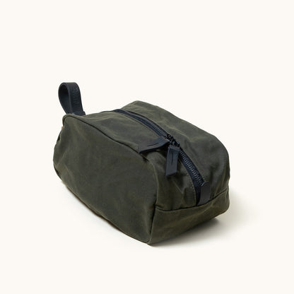 Dark green waxed canvas Drifter Dopp Kit with a black zipper, two leather pull tabs, and a leather carry loop, shown at an angle.