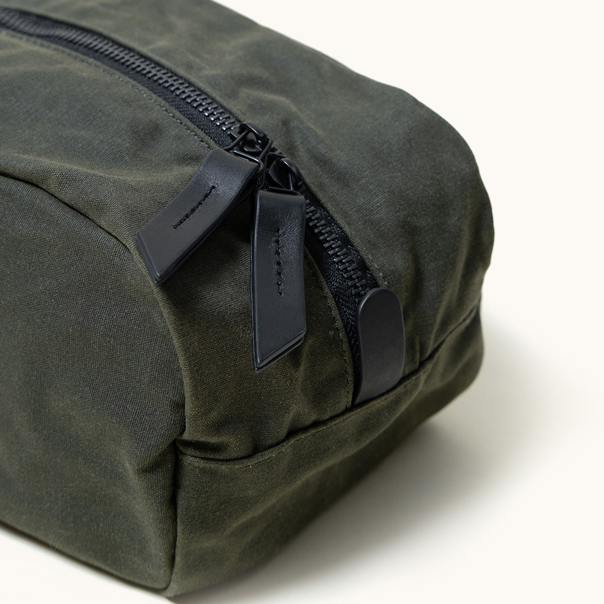 Juniper green waxed canvas Dopp Kit with black zippers and leather pull tabs, shown in a close-up, angled view.