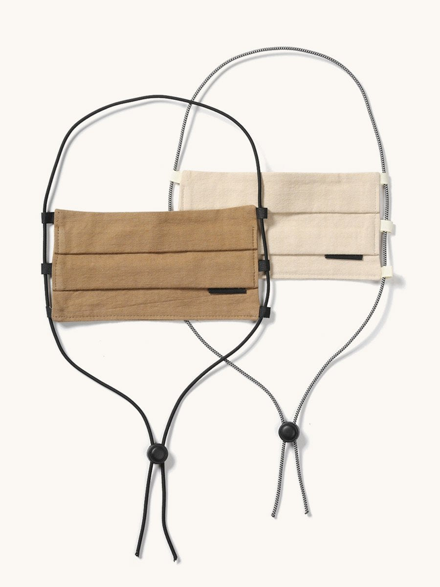 Two pleated fabric face coverings, one tan and one cream, with adjustable black and patterned lanyards.