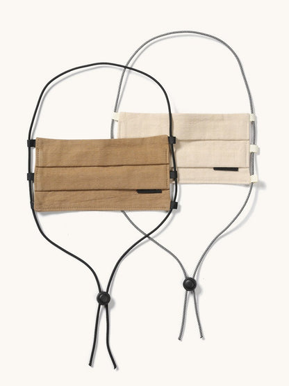Two pleated fabric face coverings, one tan and one cream, with adjustable black and patterned lanyards.