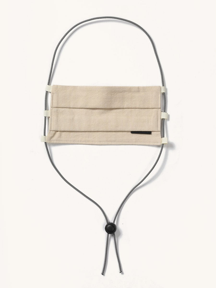 Beige pleated fabric face covering with a small black tag and black and white striped lanyard featuring a cord lock.