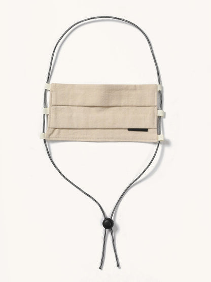 Beige pleated fabric face covering with a small black tag and black and white striped lanyard featuring a cord lock.