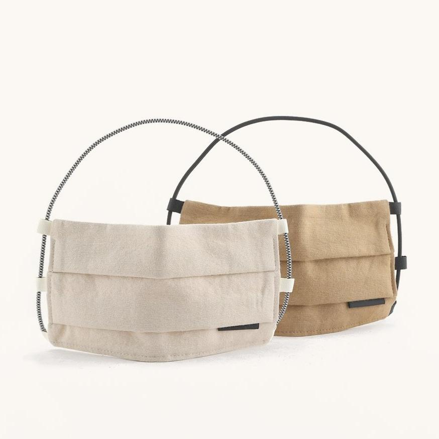 Two soft pleated fabric face coverings, one light beige with a striped cord and one tan, suspended by black handles.
