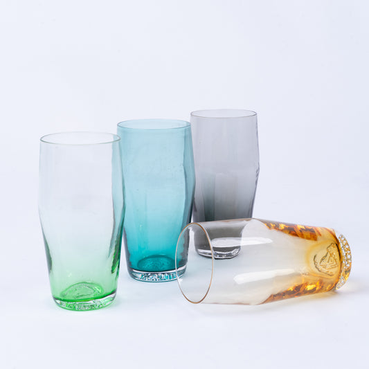 Four b grade 20 oz pint glasses in green, blue, gray, and amber glass, three standing upright and one lying on its side.
