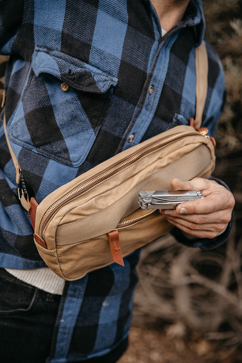 Tan waxed canvas crossbody pack with a mesh pocket and leather accents, worn by a person holding a multi-tool.