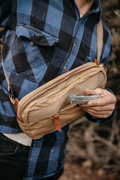 Tan waxed canvas crossbody pack with a mesh pocket and leather accents, worn by a person holding a multi-tool.