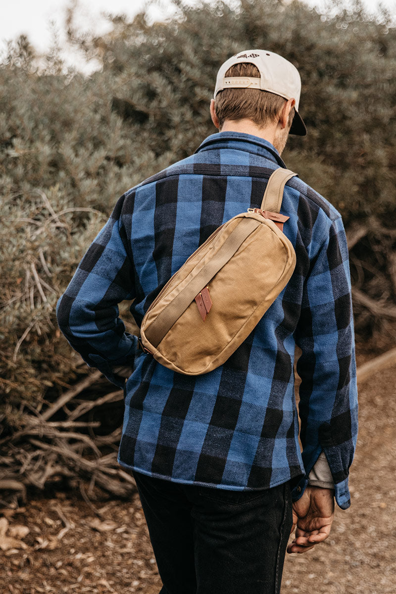 Utility Tan Waxed Canvas Canyon Crossbody Pack worn across a man's back over a blue and black plaid shirt in an outdoor setting.
