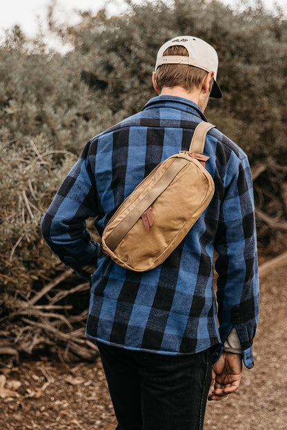 Utility Tan Waxed Canvas Canyon Crossbody Pack worn across a man's back over a blue and black plaid shirt in an outdoor setting.