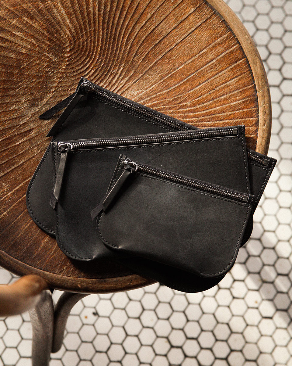 Three black leather Universal Zip Pouches, stacked and varying in size, on a wooden chair above hexagonal tiles.