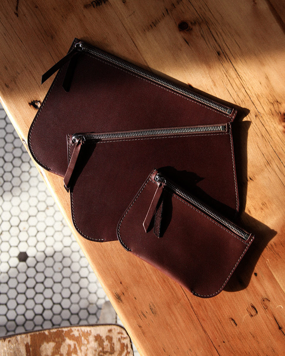 Three burgundy leather Universal Zip Pouches of varying sizes, with metal zippers and pull tabs, on a wooden table.