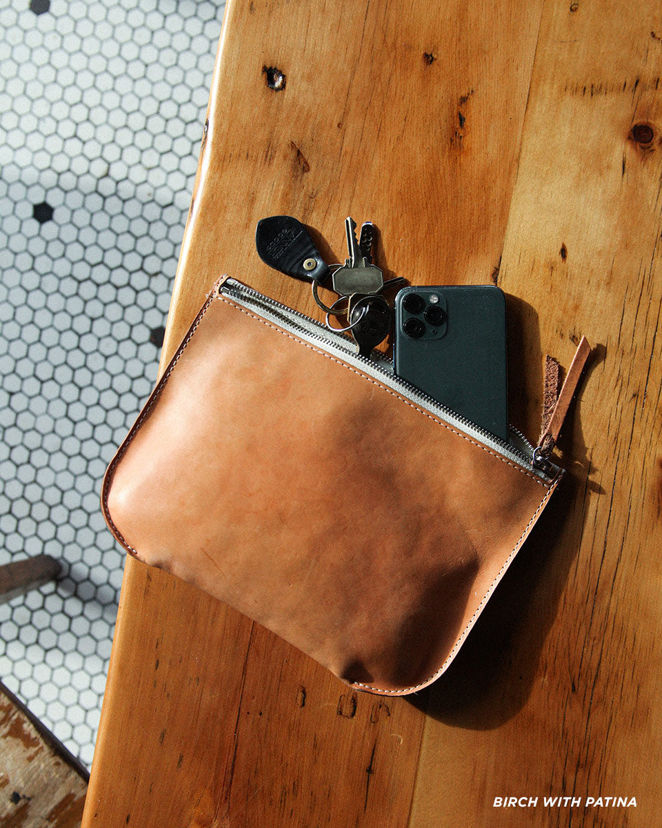 Tan leather universal zip pouch, partially unzipped, holding a dark smartphone and keys, on a wooden surface.