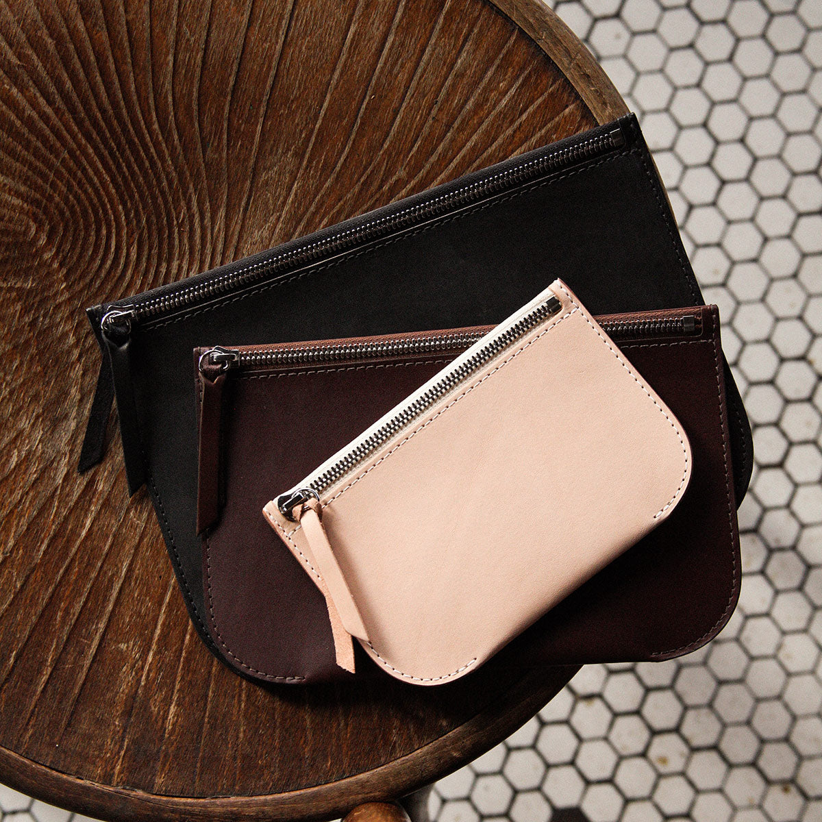 Stacked Universal Zip Pouches in natural, dark brown, and black leather on a rustic wooden table.