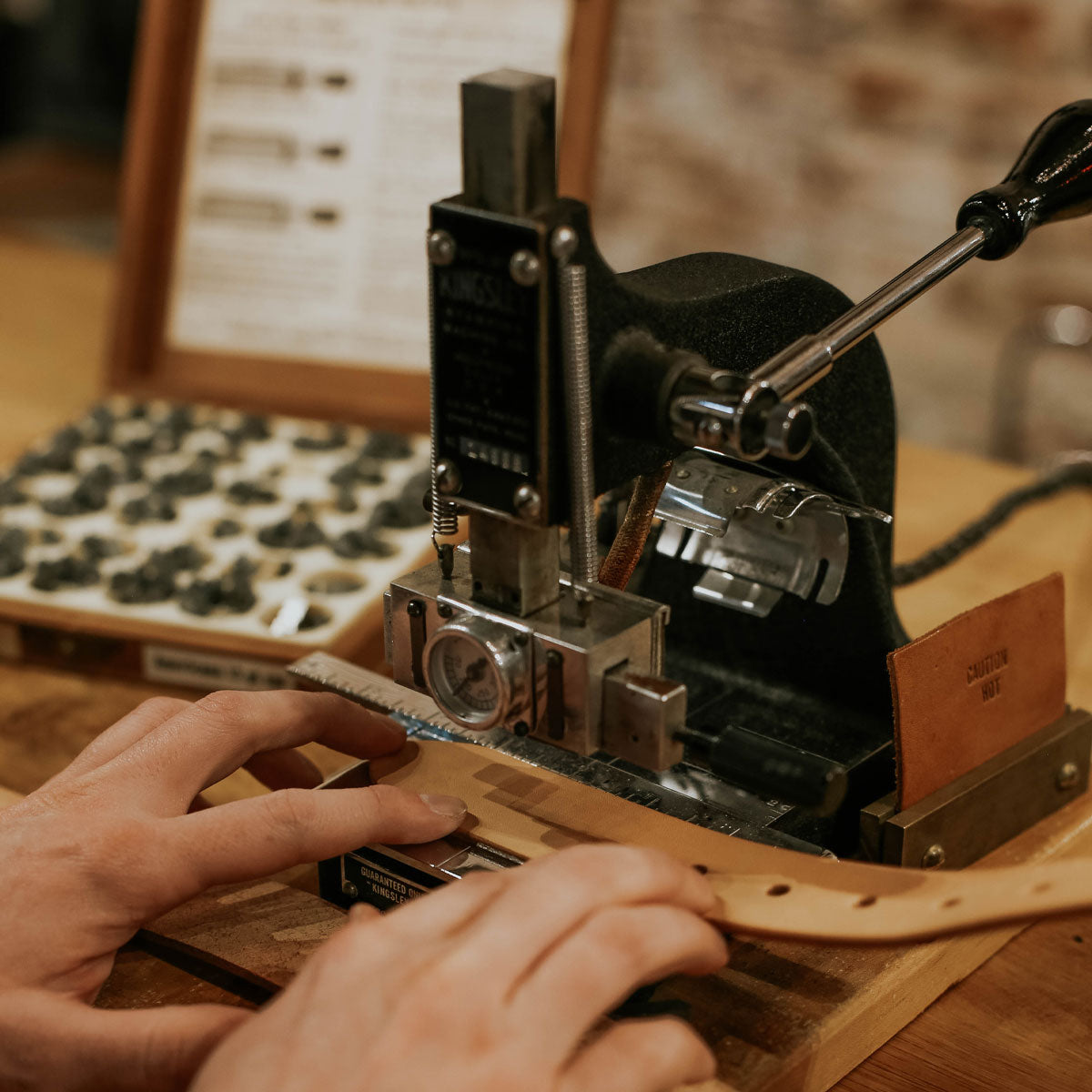Monogram hot-stamped onto a light brown leather belt by hands using a black machine with metal characters.