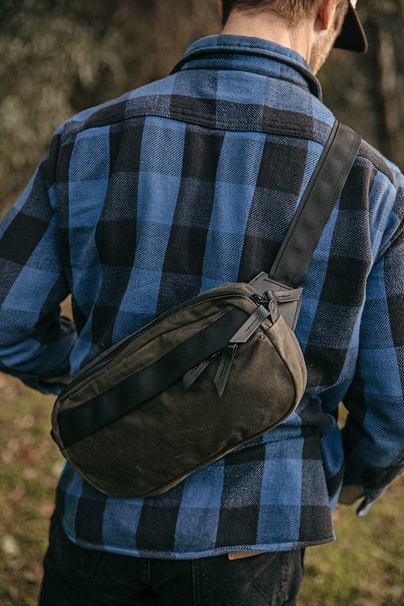 Juniper green waxed canvas crossbody pack with dark accents, worn across a person's back over a blue and black plaid shirt.