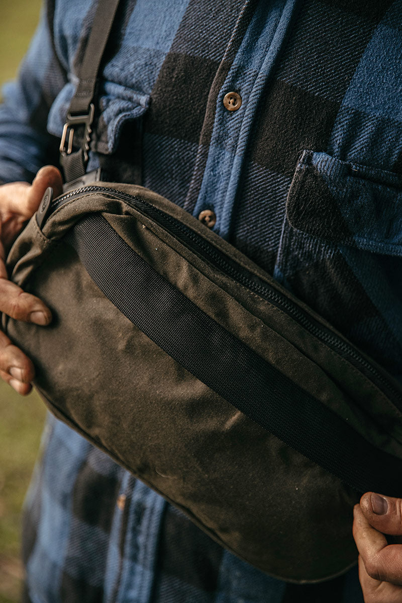 Juniper green waxed canvas crossbody pack with black zipper and strap, held by hands over a plaid shirt.