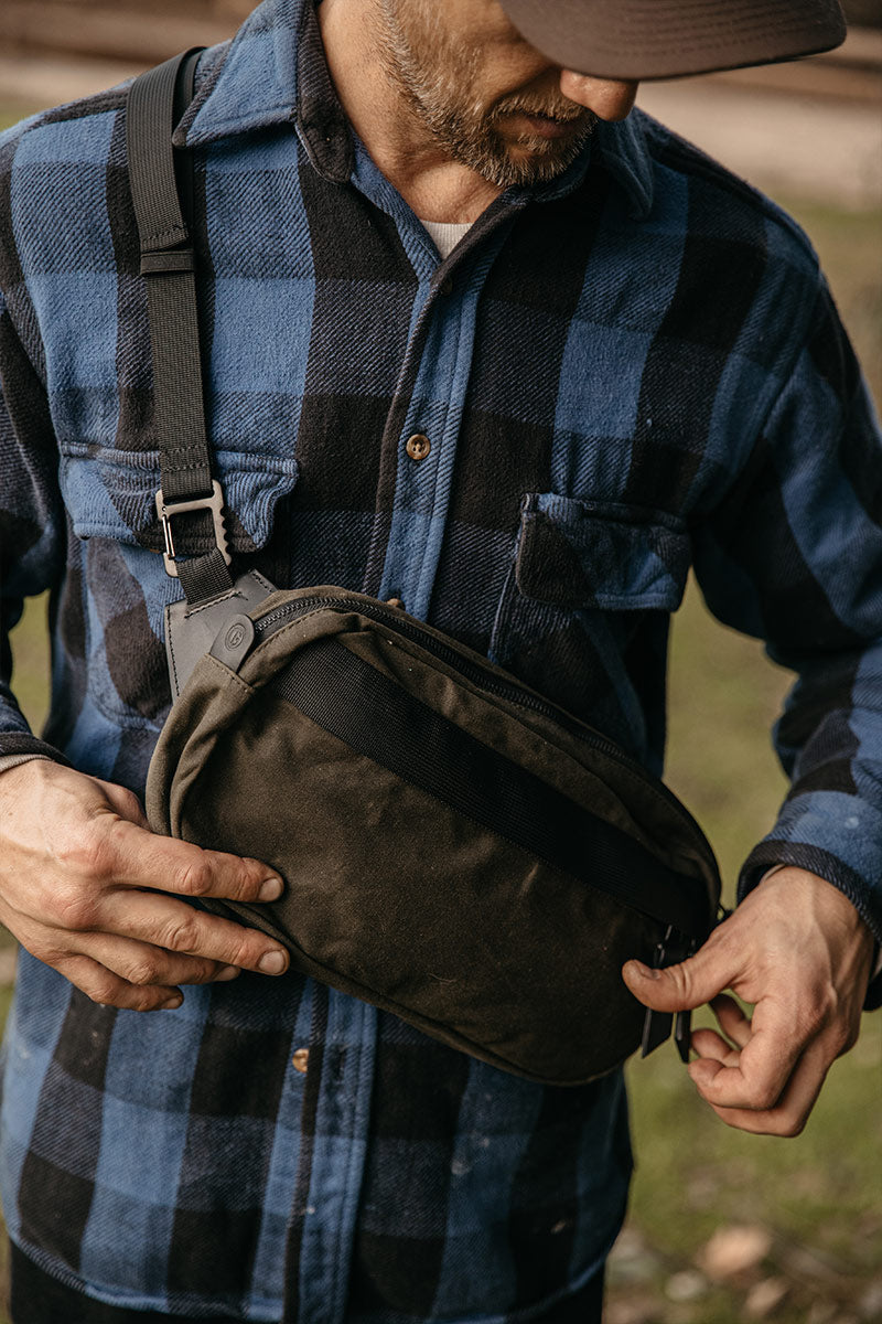 Juniper green waxed canvas crossbody pack, held by a person in a blue and black plaid shirt.