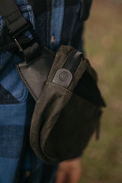 Juniper Green waxed canvas Canyon Crossbody Pack with a black strap, leather trim, and logo zipper pull, worn on a blue plaid shirt.