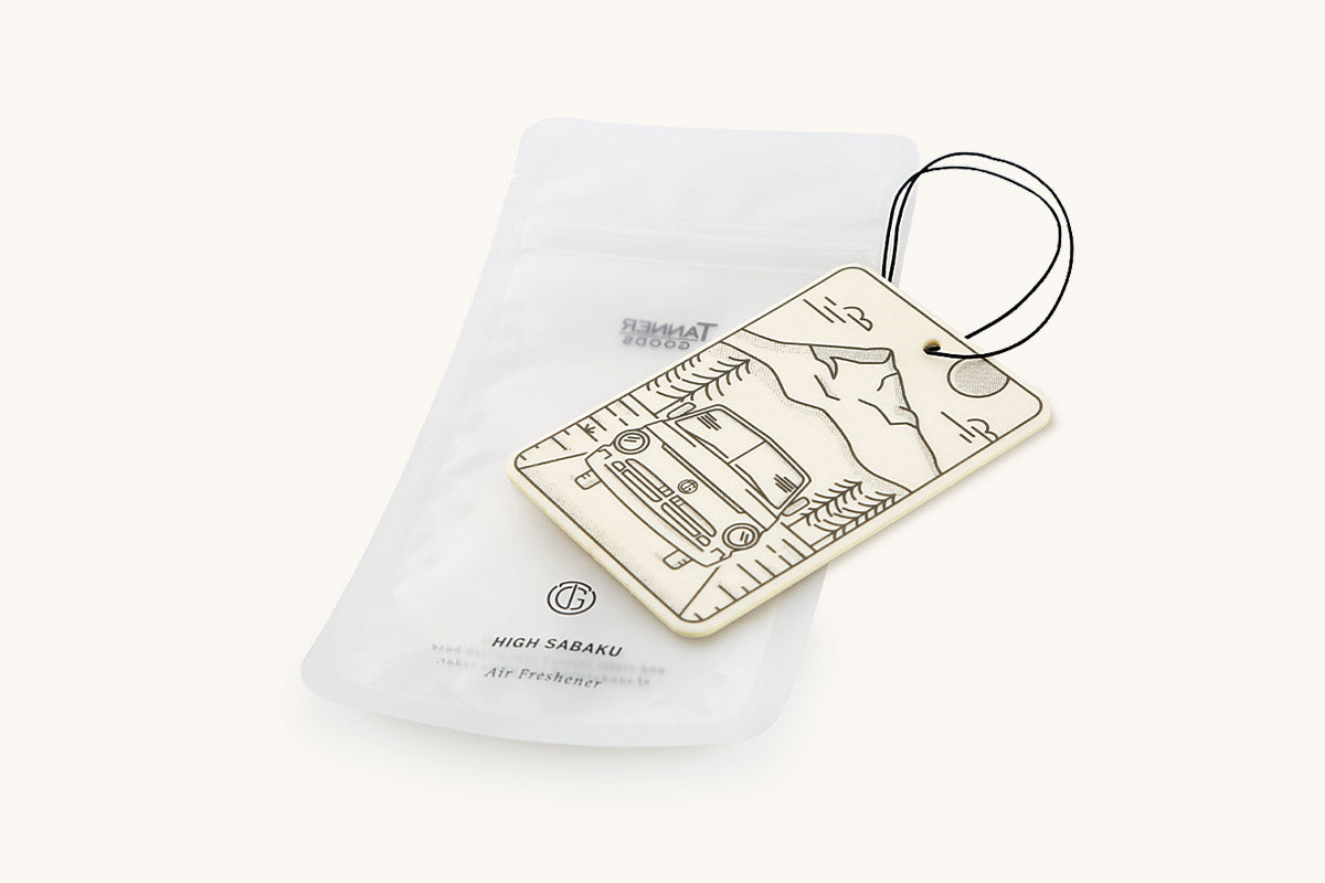 Off-white rectangular air freshener by Tanner Goods, featuring a mountain and car illustration, with a black string loop and translucent pouch.