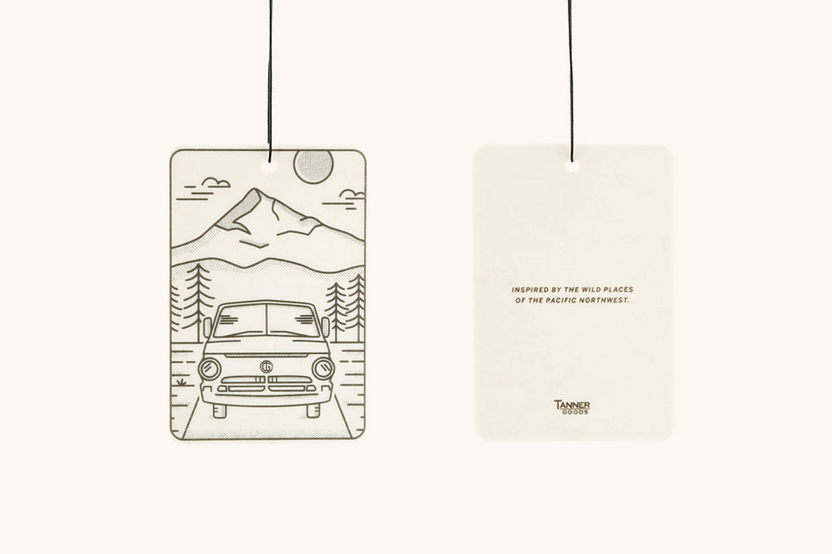 White rectangular air freshener by Tanner Goods, displaying a camper van and mountains illustration on the front, and text on the back, both hanging.