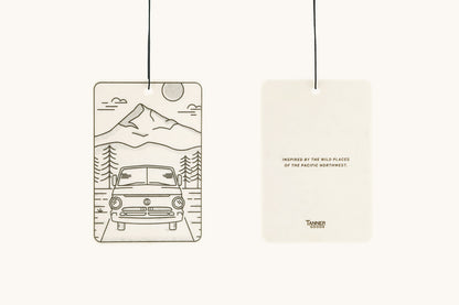 White rectangular air freshener by Tanner Goods, displaying a camper van and mountains illustration on the front, and text on the back, both hanging.