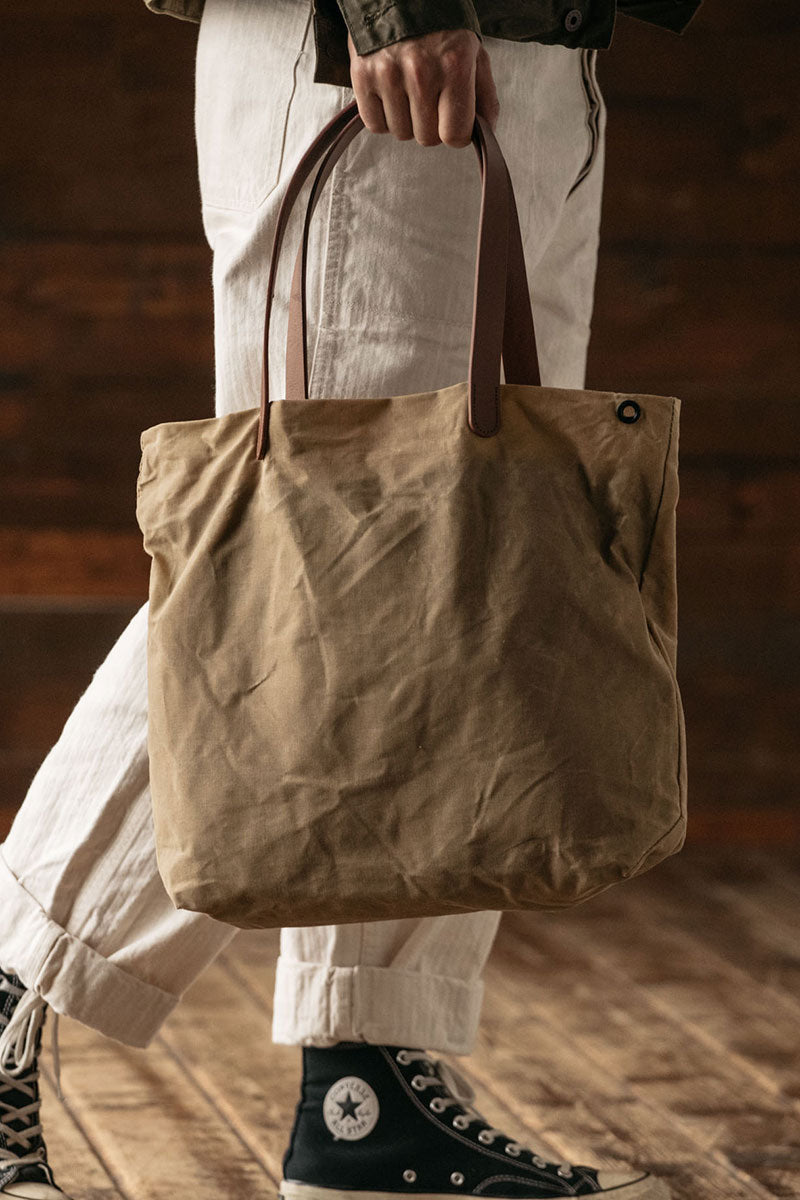 Utility tan waxed canvas simple tote bag with brown leather handles and a dark metal eyelet, carried by a person wearing light cuffed pants and dark high-top sneakers.