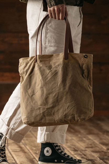 Utility tan waxed canvas simple tote bag with brown leather handles and a dark metal eyelet, carried by a person wearing light cuffed pants and dark high-top sneakers.