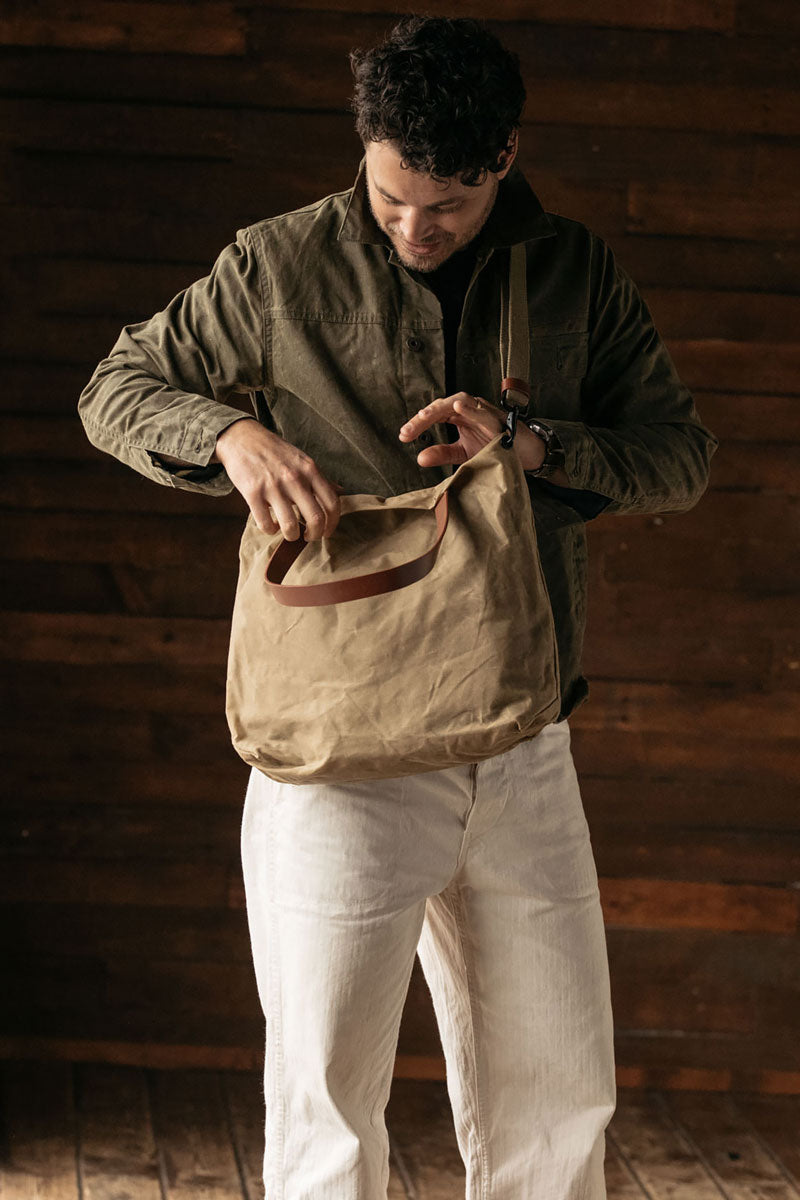 Waxed canvas tote bag in utility tan with a brown leather strap, held open by a man in a dark green jacket.