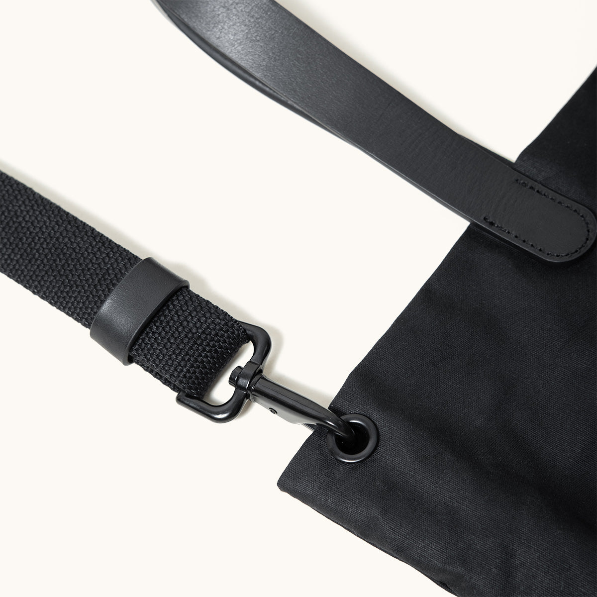 Black waxed canvas tote bag featuring a black webbing strap, leather accents, and a metal swivel hook.