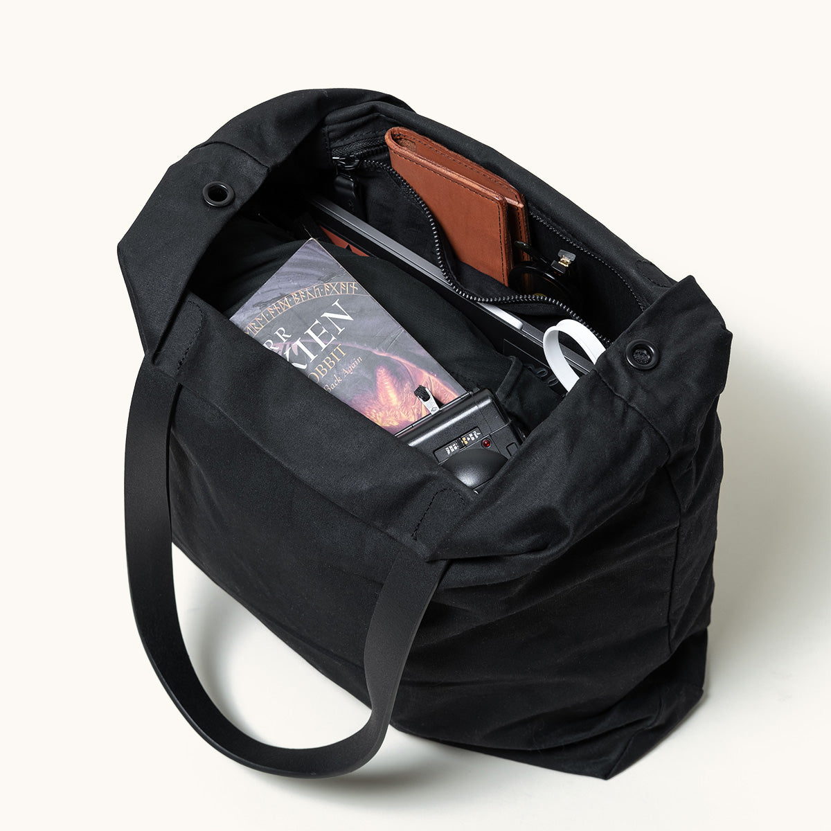 Black waxed canvas Simple Tote, open and filled with a brown wallet, The Hobbit book, and a camera, angled from above.