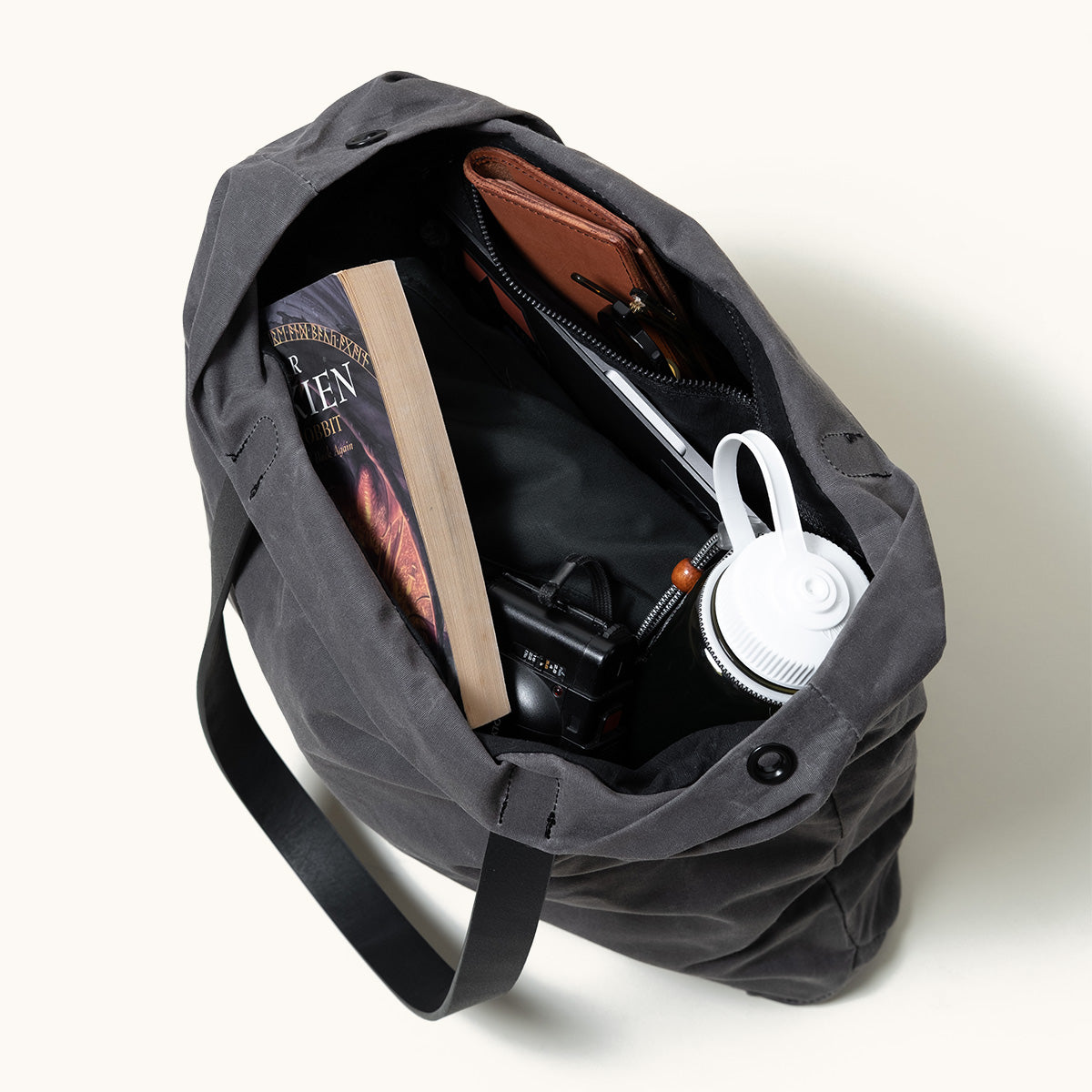 Slate grey waxed canvas tote bag, shown open from above, filled with a book, wallet, keys, camera, and water bottle.