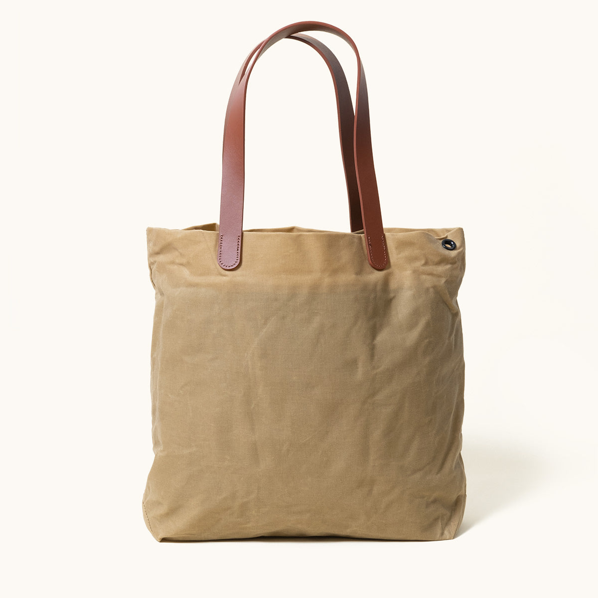 Utility Tan waxed canvas tote bag with brown leather handles and a dark metal eyelet, standing upright.
