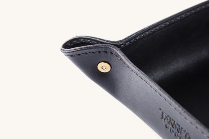 Angled view of a black leather valet tray with a brass corner snap, visible stitching, and debossed text.